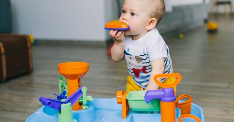 Adorable toddler chewing on toy piece while playing indoors with bright colored plastic toy. - King Squishy