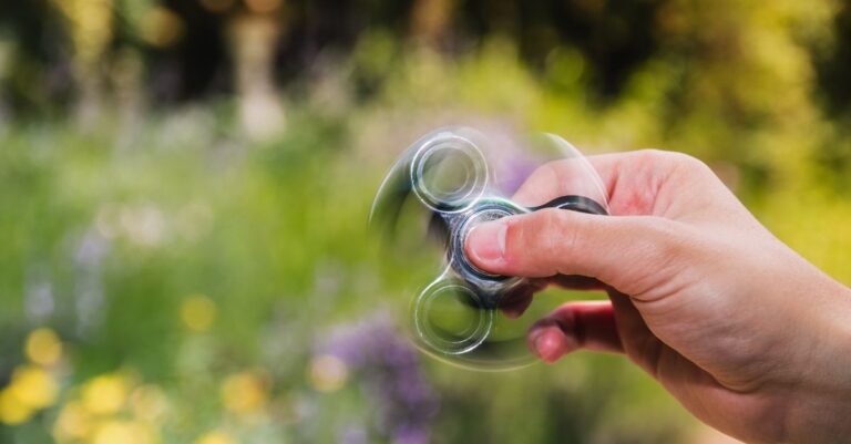Close-up of a hand spinning a fidget spinner outdoors with a blurred natural background. - King Squishy
