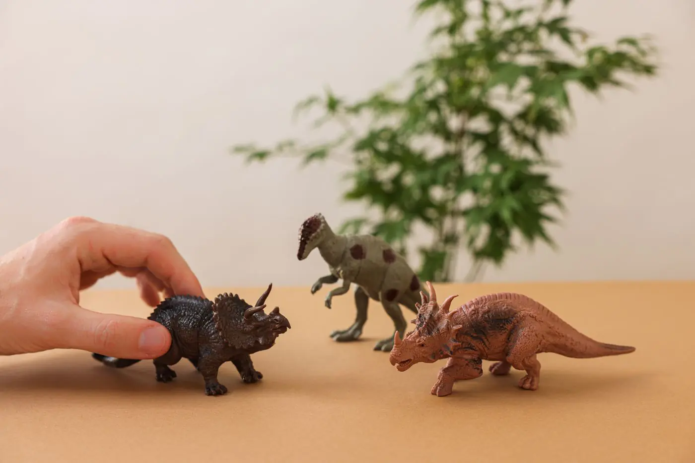 Hand arranging dinosaur figurines on a brown surface with greenery background. - King Squishy