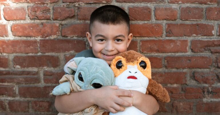 A cheerful child hugs Grogu and Porg plush toys against a rustic brick wall background. - King Squis - King Squishy