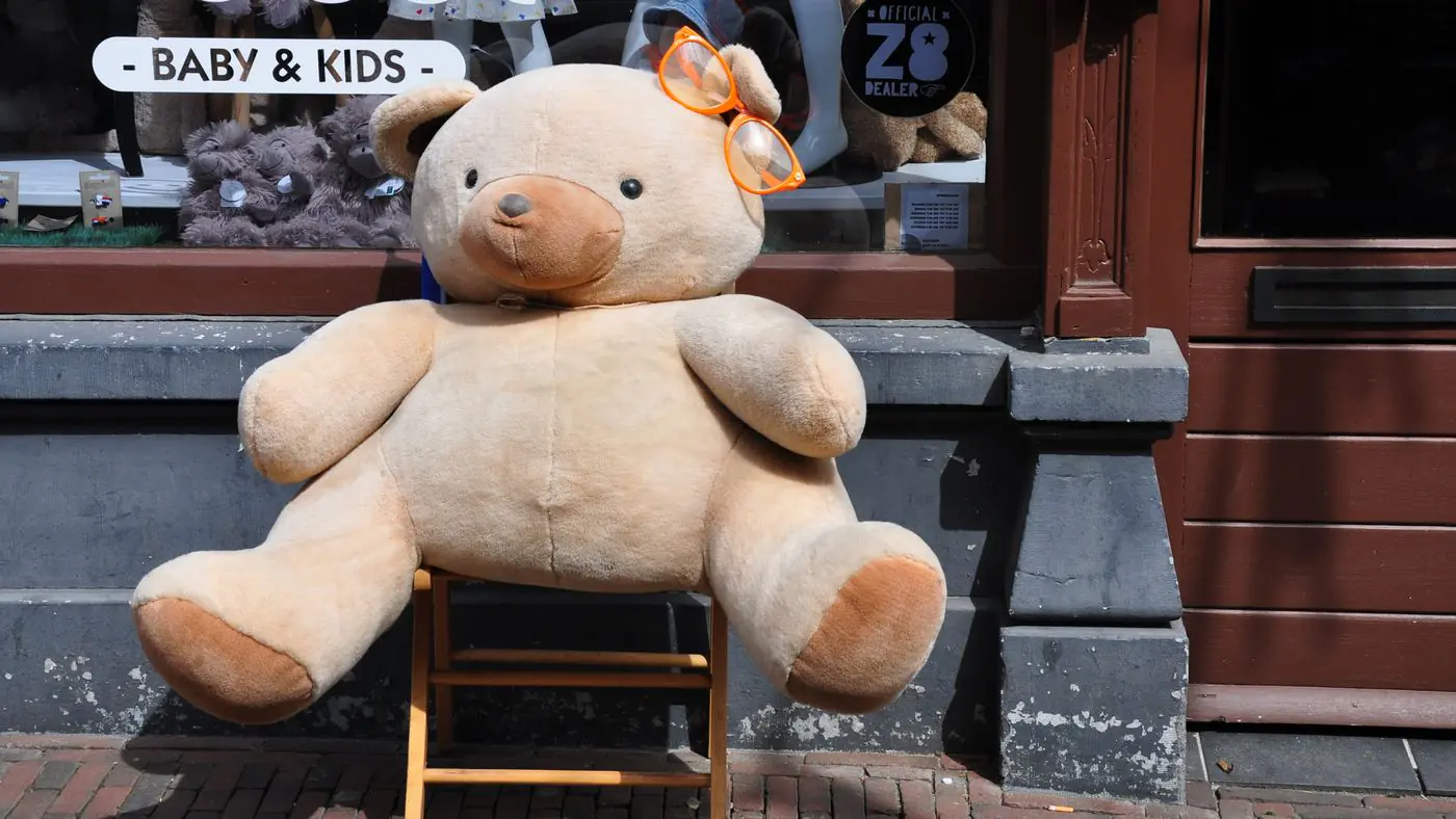 주황색 안경을 쓴 거대한 테디 베어가 완구점 진열창 앞에 놓여 있습니다. - King Squishy
