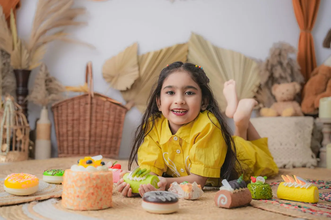 A cheerful girl in a yellow dress surrounded by colorful cakes indoors.