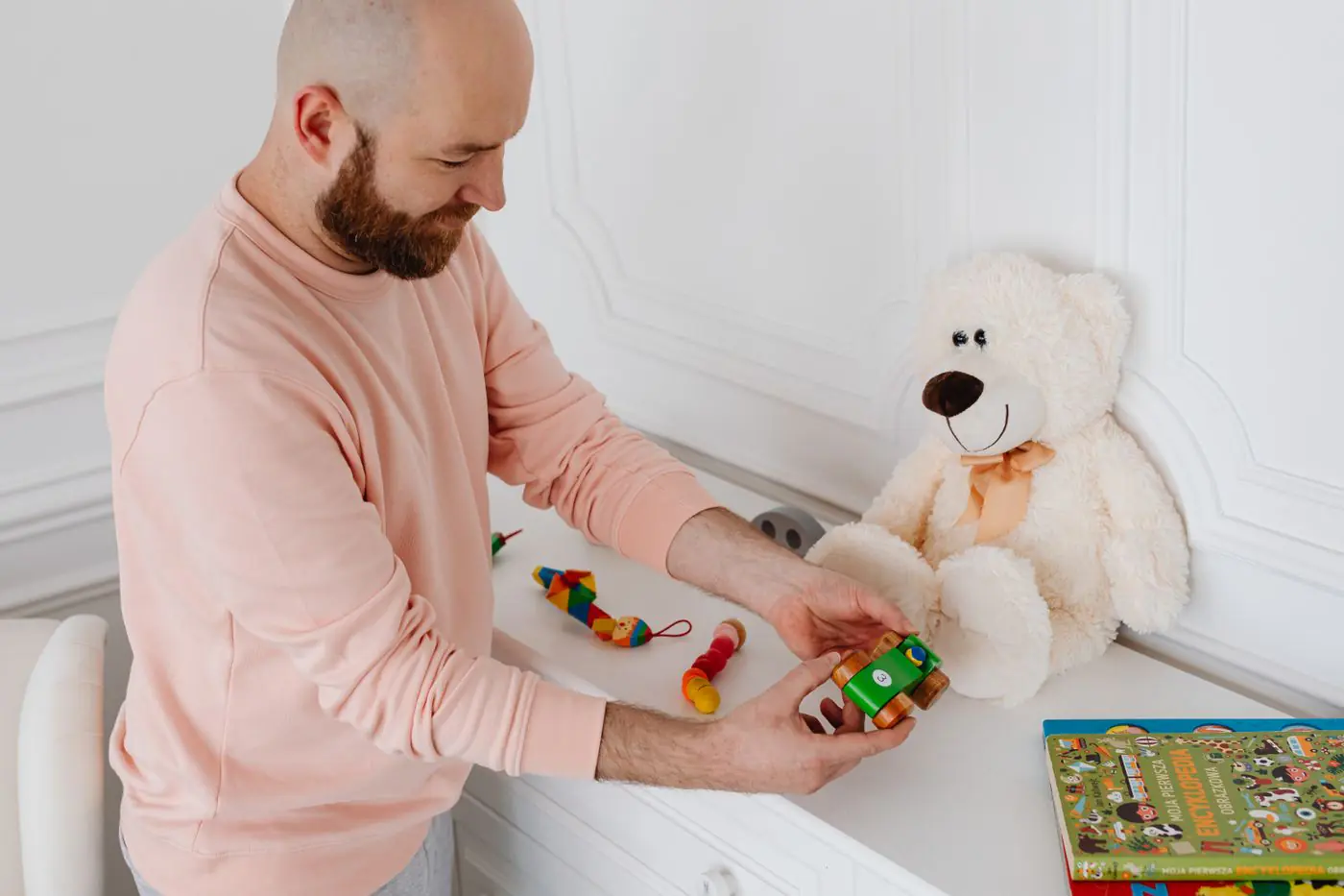 A man in a pink sweater holding a wooden toy indoors with a plush toy nearby.