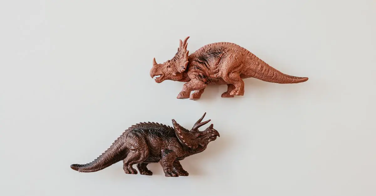 Due modelli dettagliati di giocattoli dinosauri, inclusi un Triceratopo, su una superficie bianca. - King Squishy