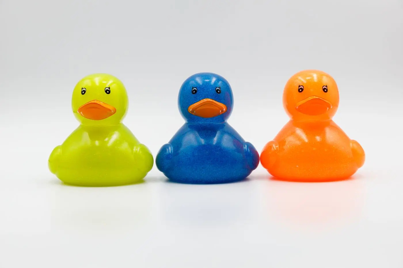Tiga bebek karet berwarna-warni hijau, biru, dan oranye di latar putih. - King Squishy