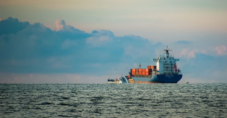 A cargo ship with containers navigates the vast open sea at dusk under a colorful sky. - King Squishy