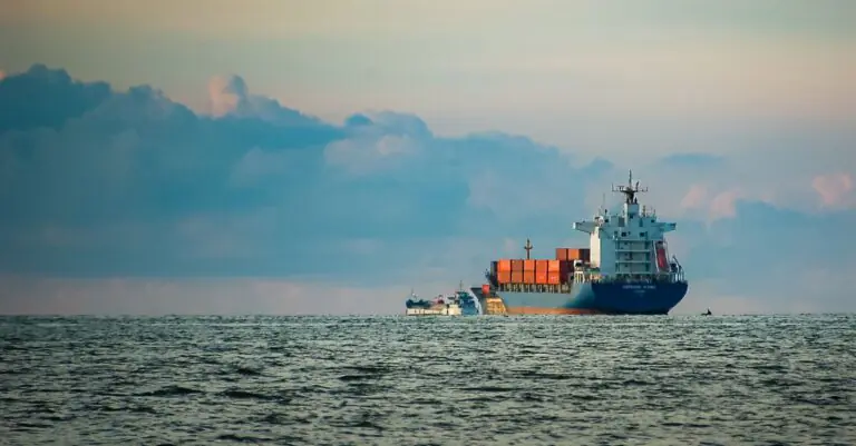 A cargo ship with containers navigates the vast open sea at dusk under a colorful sky. - King Squishy