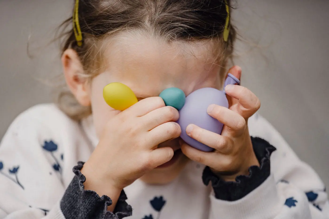 Crop anonymous shy kid in ornamental apparel with barrettes covering eyes with various balloons in daylight - King Squishy