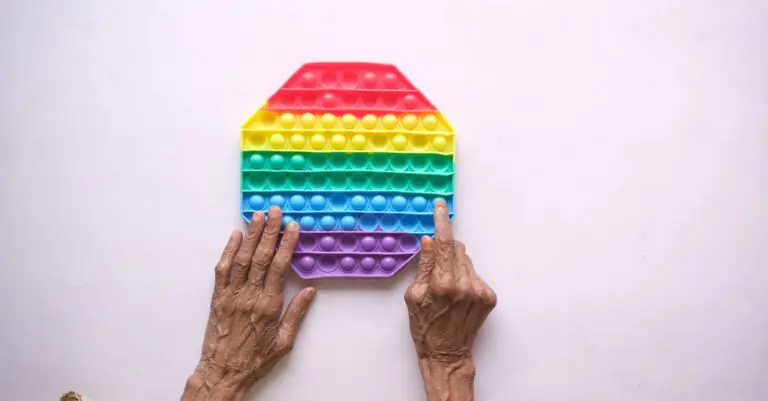 Close-up of elderly hands on a rainbow silicon push popper toy on white background. - King Squishy
