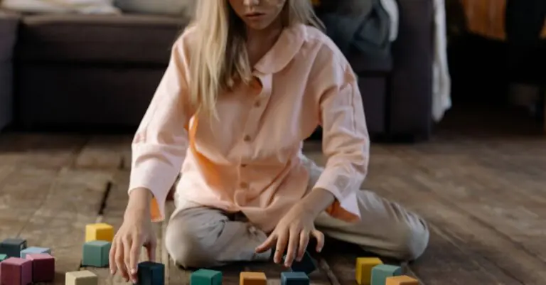 A young girl sits indoors playing with colorful wooden cubes on the floor. - King Squishy