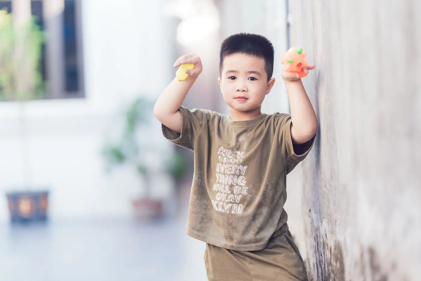 A cheerful child holding colorful balls while leaning on a wall outdoors.